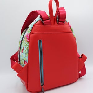 Puede incluir: Una mochila de cuero roja con un panel de tela estampado floral en la parte delantera. La mochila tiene un bolsillo con cremallera en la parte posterior y correas ajustables.