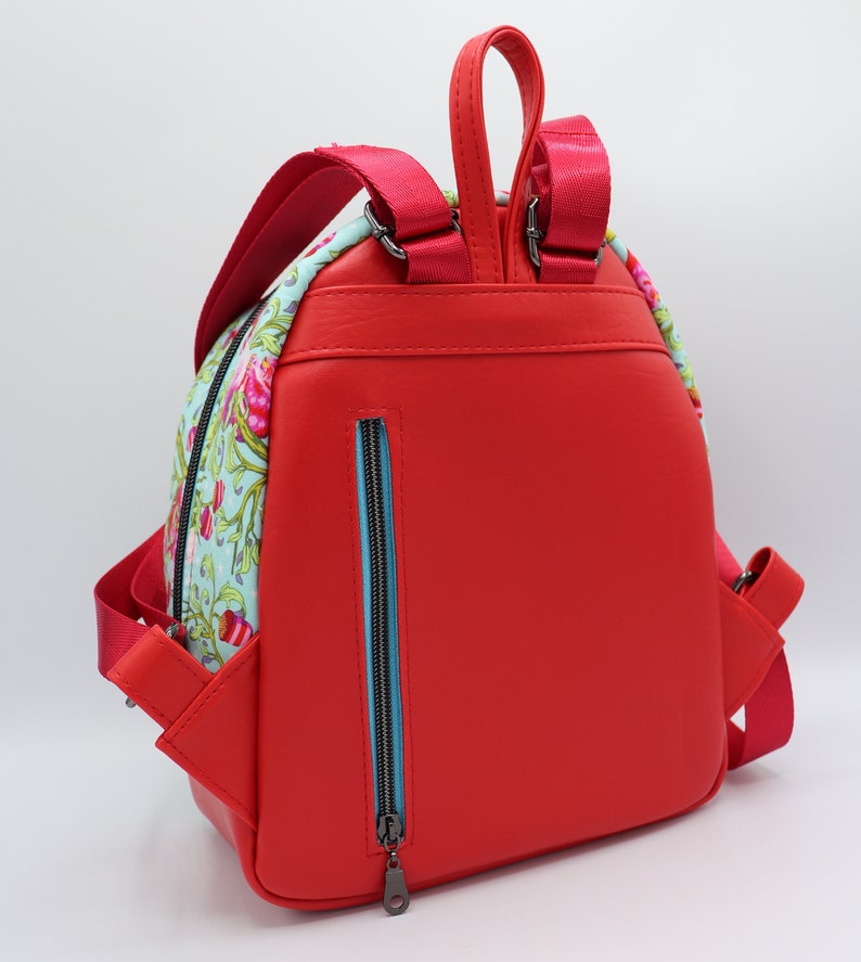 Puede incluir: Una mochila de cuero roja con un panel de tela estampado floral en la parte delantera. La mochila tiene un bolsillo con cremallera en la parte posterior y correas ajustables.