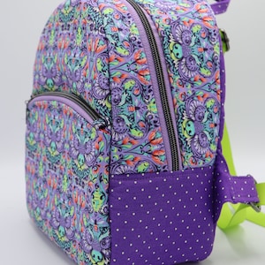 Puede incluir: Mochila de color morado y verde con estampado y fondo morado de lunares. La mochila tiene cierre de cremallera y una correa verde.