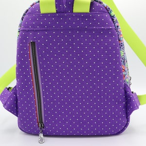 Puede incluir: Mochila de lunares morados con una correa verde lima y un bolsillo con cremallera en la parte posterior.