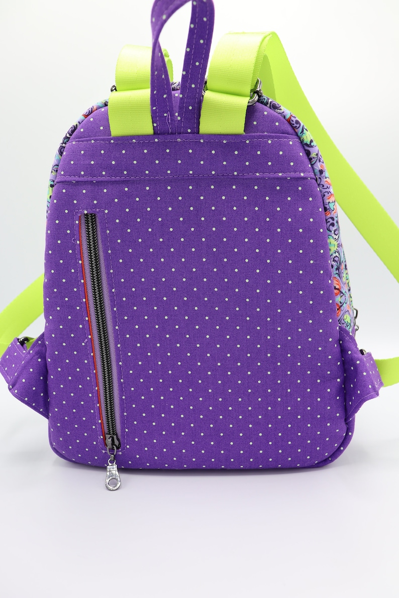Puede incluir: Mochila de lunares morados con una correa verde lima y un bolsillo con cremallera en la parte posterior.