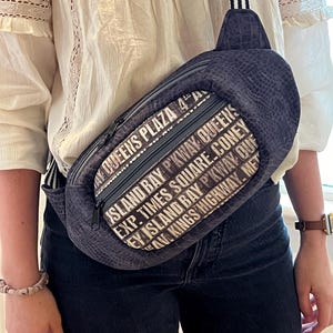 Könnte beinhalten: Eine schwarze Bauchtasche mit einem weißen Reißverschluss und einem weißen Aufdruck mit der Aufschrift "Queens Plaza 4A, Island Bay P'Kway Queens, Exp. Times Square Coney, Ey Island Bay P'Kway Ouf, Ave Kings Highway. Met"