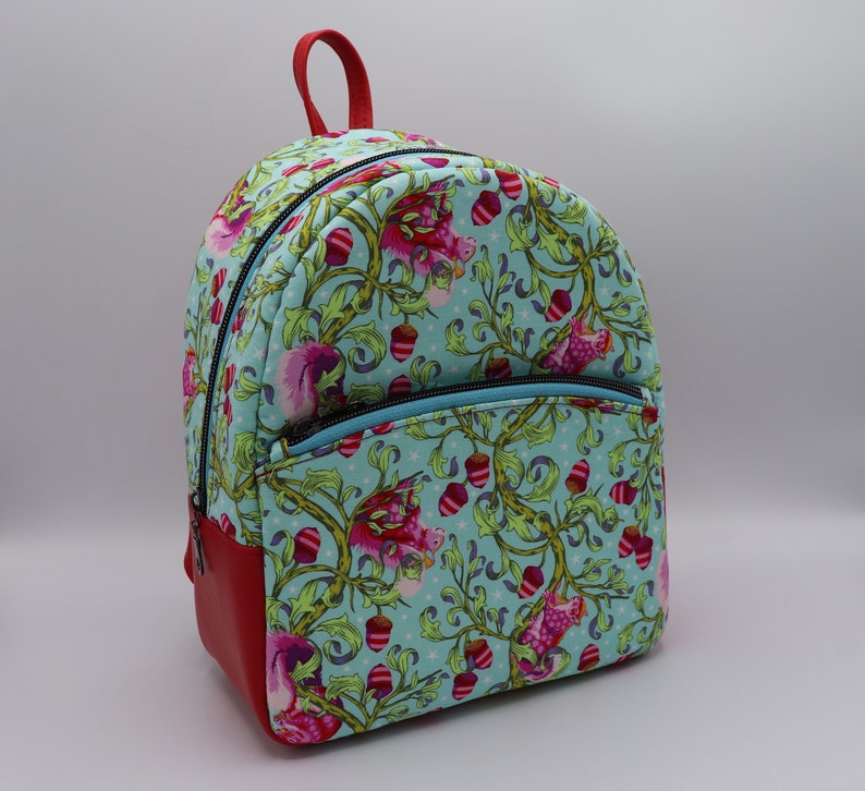 Puede incluir: Una mochila azul y roja con un estampado floral con ardillas rosas y bellotas. La mochila tiene un bolsillo con cremallera en la parte delantera.