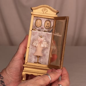 Op de afbeelding: Een miniatuur goudkleurige vitrinekast met een open glazen deur. Binnenin is een miniatuur paspop met een met kant afgewerkte top zichtbaar. De kast bevat ook miniatuur accessoires en wordt door handen vastgehouden.