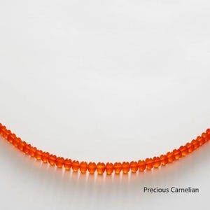 May include: A strand of orange carnelian beads, curved against a white background. The beads are small, round, and translucent, with a vibrant, warm color. The text "Precious Carnelian" is in the lower right corner.