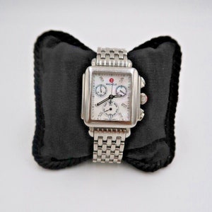 May include: A silver-toned Michele watch with a square face and a mother-of-pearl dial. The watch has a chronograph function and a date window. The watch is on a silver-toned metal bracelet.