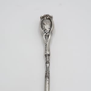 May include: A silver shoe horn with a floral design on the handle. The handle is ornate and the hook is simple.