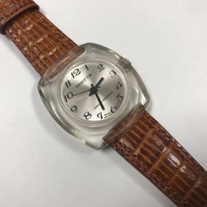Peut inclure: Une montre-bracelet de forme carr&eacute;e avec un bo&icirc;tier en plastique transparent et un bracelet en cuir marron. Le cadran de la montre est blanc avec des chiffres noirs et les mots "PRINCETON" et "17 JEWELS" imprim&eacute;s dessus.