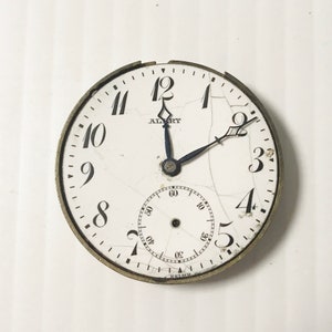 Op de afbeelding: Een wit zakhorloge met zwarte Romeinse cijfers en een zwarte secondewijzer. Het horloge heeft een gebarsten wijzerplaat met de tekst "ALBERT" en "SIMS".