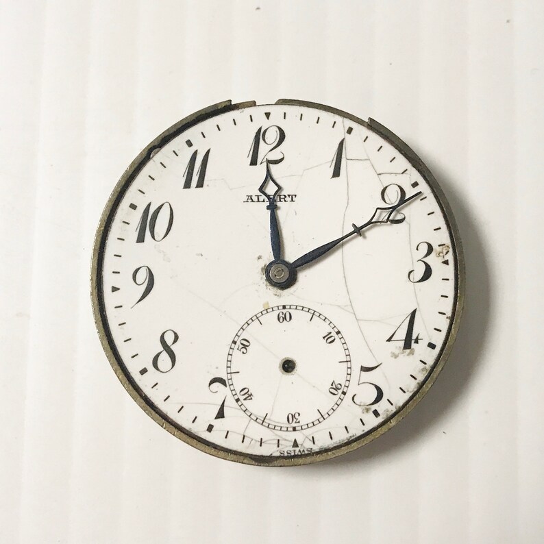 Op de afbeelding: Een wit zakhorloge met zwarte Romeinse cijfers en een zwarte secondewijzer. Het horloge heeft een gebarsten wijzerplaat met de tekst "ALBERT" en "SIMS".