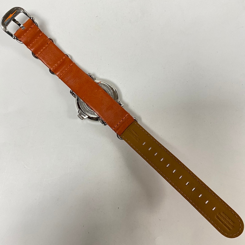May include: A wristwatch with a silver-colored face and a bright orange leather band. The band is attached to the watch with silver-colored metal rings. The watch band is approximately 9 inches long.