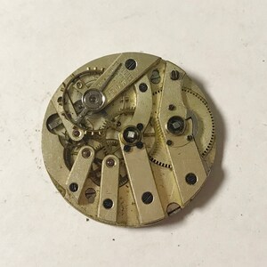Puede incluir: Primer plano de un movimiento de reloj plateado con engranajes y palancas intrincados. El movimiento tiene una forma circular y un di&aacute;metro de aproximadamente 6,5 cm.