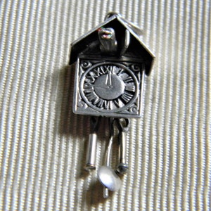 May include: Silver charm in the shape of a cuckoo clock with a black face and Roman numerals. The clock has a small bird on top and three dangling bells.