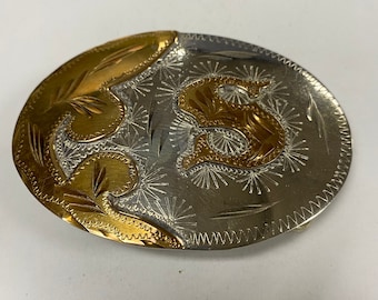 May include: A silver and gold belt buckle with an intricate design. The buckle features a stylized letter "S" in gold with a floral pattern.