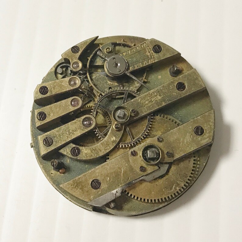 May include: Close-up of a vintage pocket watch movement with intricate gears and brass components. The watch face is missing, revealing the inner workings of the timepiece.