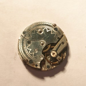 Puede incluir: Primer plano de un movimiento de reloj de bolsillo plateado con engranajes intrincados y un dise&ntilde;o decorativo. La esfera del reloj es visible con el texto "15 Jewels" y "Swiss Made".
