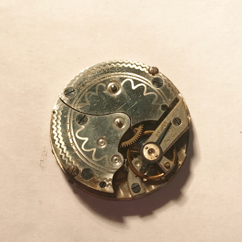 Puede incluir: Primer plano de un movimiento de reloj de bolsillo plateado con engranajes intrincados y un dise&ntilde;o decorativo. La esfera del reloj es visible con el texto "15 Jewels" y "Swiss Made".