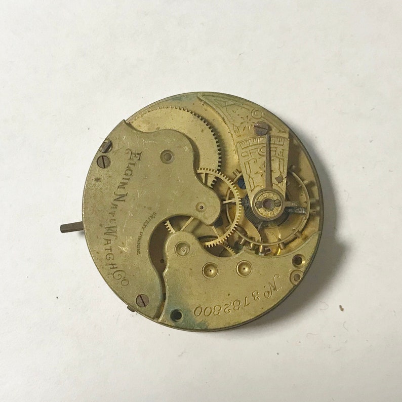 May include: A vintage Elgin National Watch Co. watch movement with visible gears and the serial number 3762800. The metal components have a gold tone, and the text "Elgin National Watch Co." is visible.