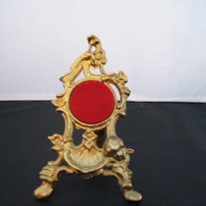 May include: A gold-colored metal stand with an ornate design. The stand has a round, red velvet top and three legs.