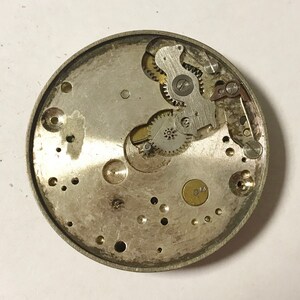 May include: Close-up view of the inner workings of a silver watch mechanism. The intricate gears and levers are visible, showcasing the complex design of the watch.