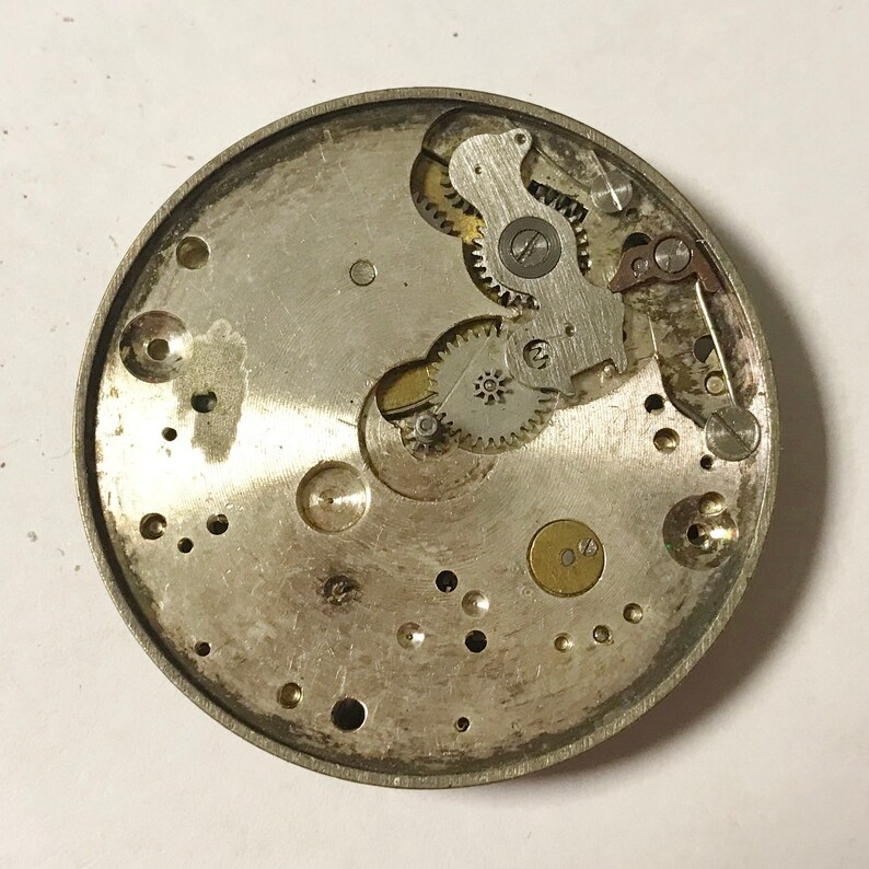 May include: Close-up view of the inner workings of a silver watch mechanism. The intricate gears and levers are visible, showcasing the complex design of the watch.