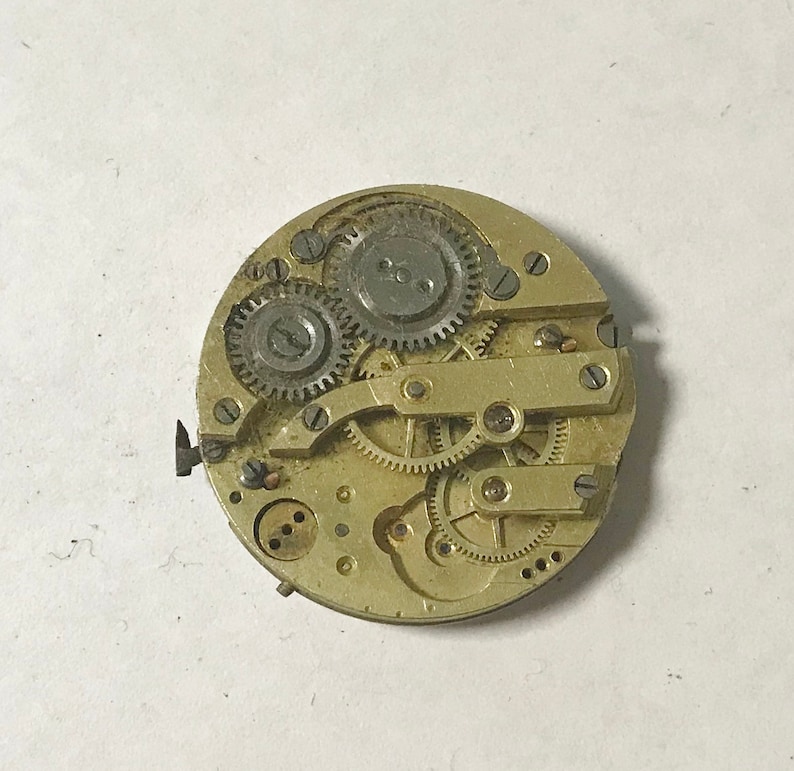 May include: Close-up of a gold-toned watch movement with multiple gears and a winding stem. The watch movement is exposed and shows the intricate workings of the watch.