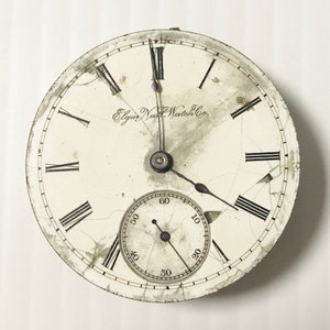 May include: A white pocket watch with Roman numeral hour markers and a smaller seconds dial. The watch face is cracked and has the text "Elgin Natl. Watch Co." printed on it.