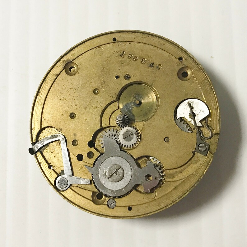 May include: A detailed view of a vintage watch movement. The circular brass plate houses numerous small gears, levers, and a balance wheel. The number "188945" is stamped on the brass. The mechanism is a complex arrangement of tiny components.