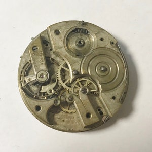 May include: Close-up of a silver pocket watch movement with intricate gears and a winding mechanism. The watch face is not visible.