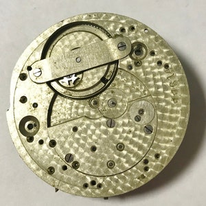 Pu&ograve; includere: Primo piano di un movimento di orologio argentato con ingranaggi intricati e una superficie testurizzata. Il movimento &egrave; etichettato "M1911".