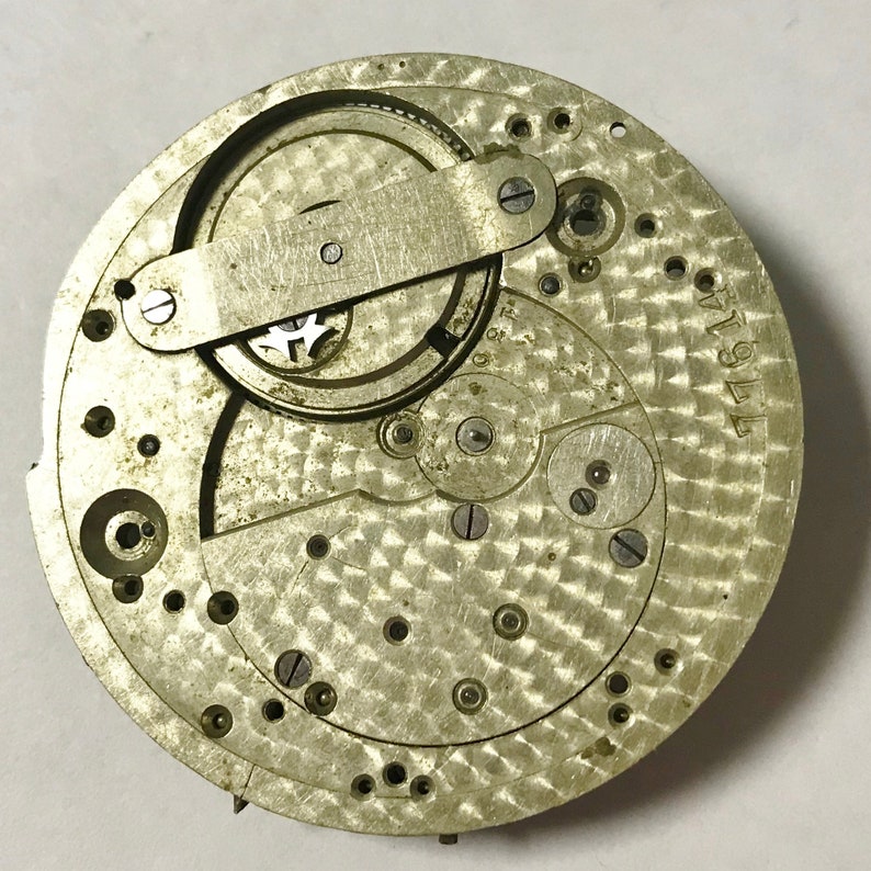 Pu&ograve; includere: Primo piano di un movimento di orologio argentato con ingranaggi intricati e una superficie testurizzata. Il movimento &egrave; etichettato "M1911".