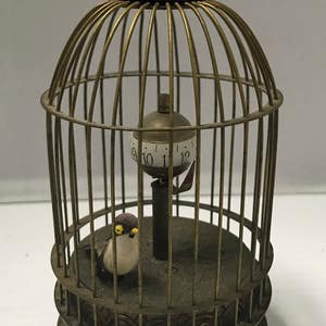 May include: A brass birdcage clock with a small bird figurine inside. The clock face is visible through the cage and has Roman numerals. The cage has a handle on top.