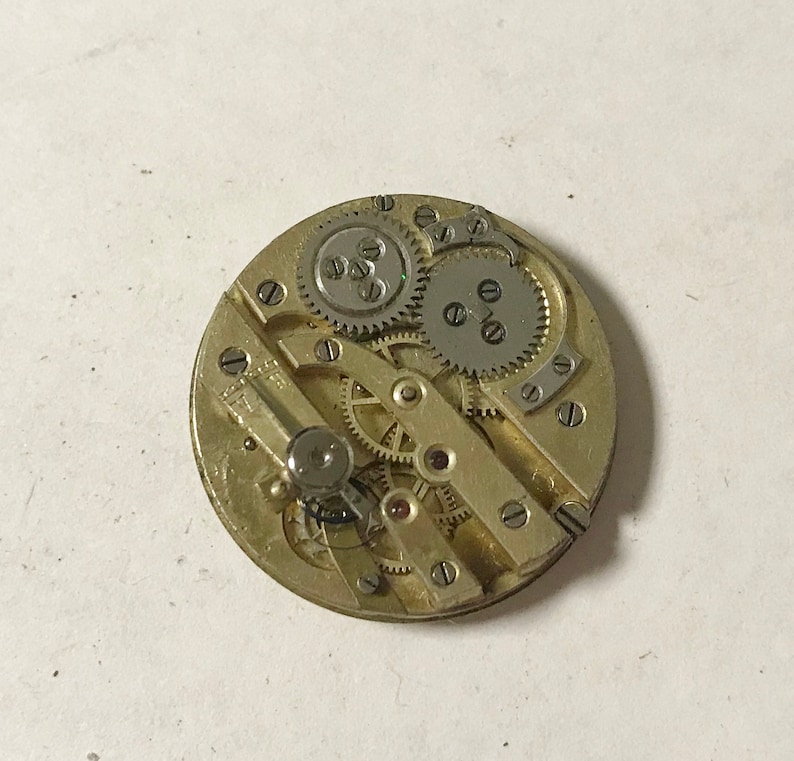 Puede incluir: Primer plano de un movimiento de reloj de tono dorado con engranajes intrincados y un v&aacute;stago de cuerda de tono plateado. El movimiento est&aacute; expuesto y visible, mostrando el funcionamiento interno del reloj.