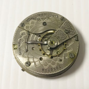 Op de afbeelding: Close-up van een zilveren zakhorlogemechanisme met de inscriptie "Elgin Nat'l Watch Co. U.S.A." en "Seven Jewels". Het horloge heeft een decoratief bloemmotief en het nummer "7629714" is zichtbaar.