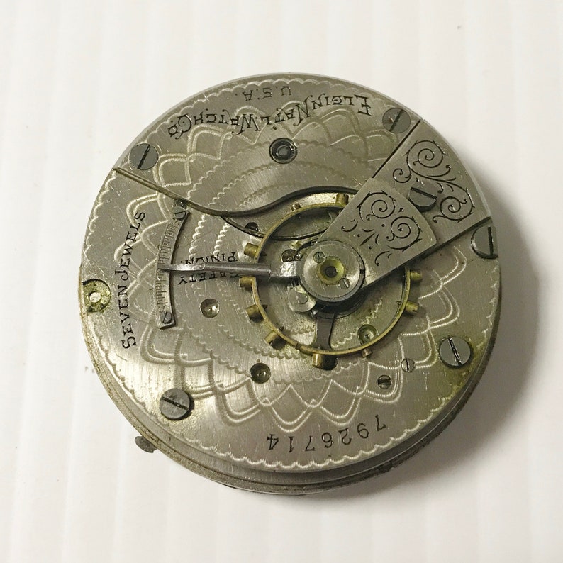 Op de afbeelding: Close-up van een zilveren zakhorlogemechanisme met de inscriptie "Elgin Nat'l Watch Co. U.S.A." en "Seven Jewels". Het horloge heeft een decoratief bloemmotief en het nummer "7629714" is zichtbaar.