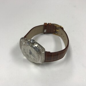 Peut inclure: Une montre-bracelet en plastique transparent avec un cadran blanc et des aiguilles noires. La montre a un bracelet en cuir marron avec un motif crocodile.