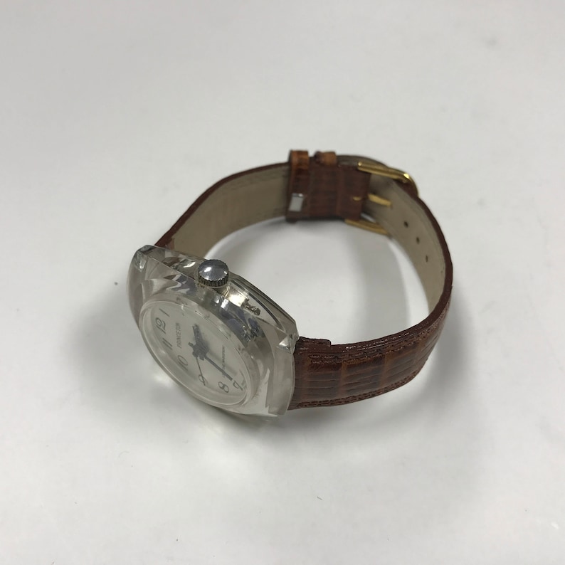 Peut inclure: Une montre-bracelet en plastique transparent avec un cadran blanc et des aiguilles noires. La montre a un bracelet en cuir marron avec un motif crocodile.