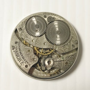 May include: Close-up of a vintage watch movement with intricate gears and mechanisms. The silver-toned components are etched with decorative patterns and text, including "WALTHAM MASS." and "SAFETY BARREL."