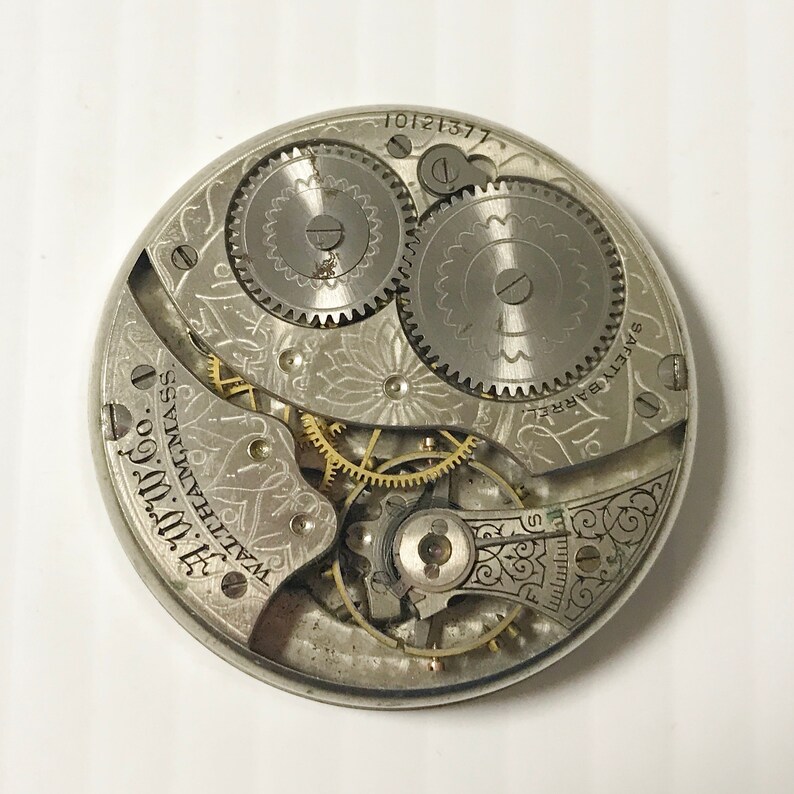 May include: Close-up of a vintage watch movement with intricate gears and mechanisms. The silver-toned components are etched with decorative patterns and text, including "WALTHAM MASS." and "SAFETY BARREL."