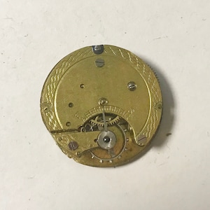May include: A gold-colored pocket watch movement with visible gears and a decorative pattern on the back.