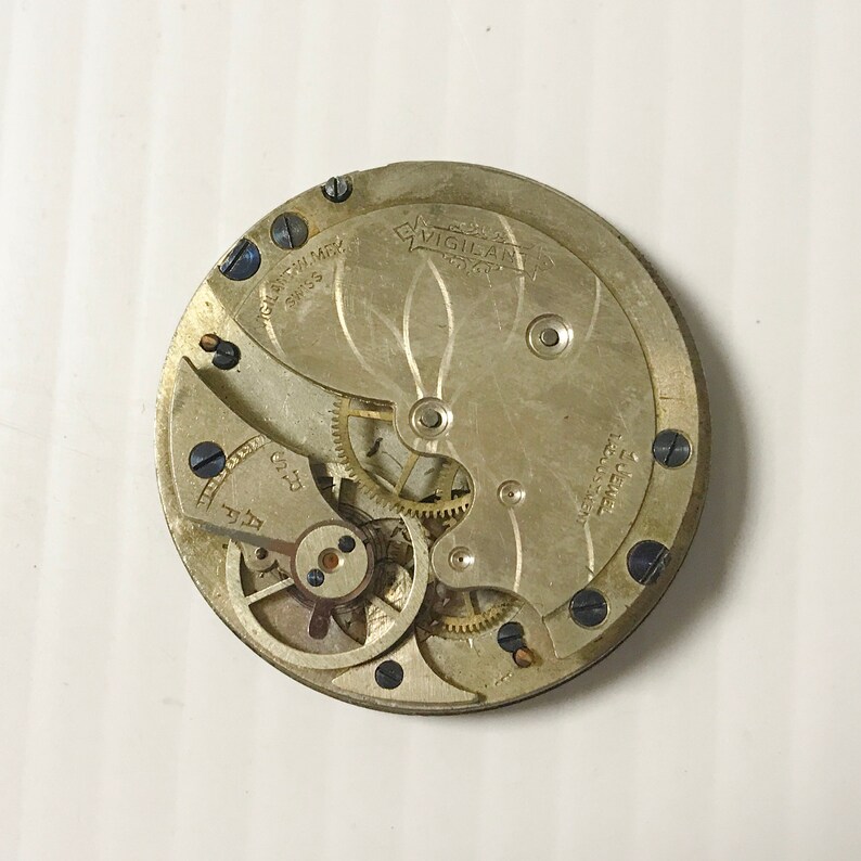 Op de afbeelding: Close-up van een zilveren zakhorloge-uurwerk met de tekst "Vigilant" en "Swiss Made" zichtbaar. Het horloge heeft een ingewikkeld tandwielsysteem en moet worden gerepareerd.
