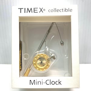 May include: A collectible Timex Mini-Clock in a clear plastic case. The clock has a gold-colored face with black numerals and hands. The clock is designed to look like a fishing rod and reel. The packaging has the words "TIMEX collectible" and "Mini-Clock".