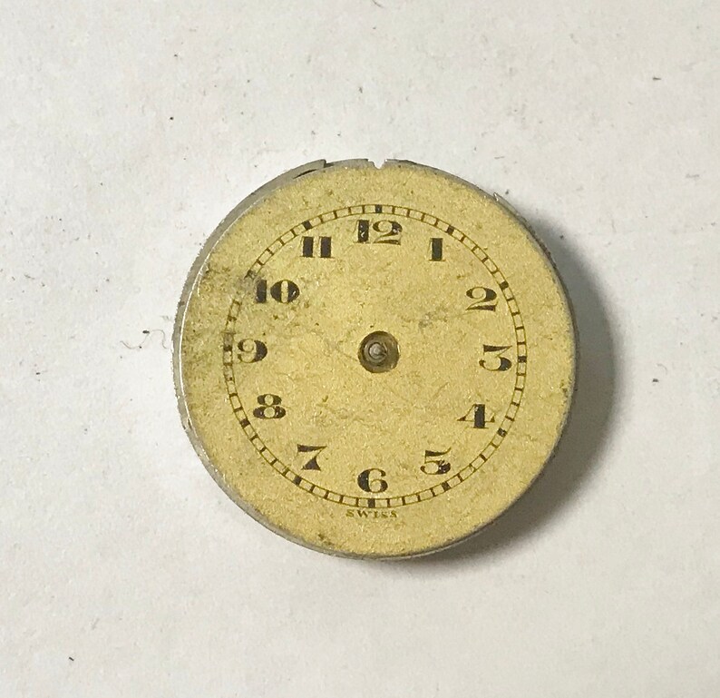 Puede incluir: Una esfera de reloj redonda de color crema con marcadores de hora en n&uacute;meros romanos negros y marcadores de minutos en n&uacute;meros ar&aacute;bigos negros. La esfera est&aacute; rodeada por un borde de metal plateado con la palabra "Swiss" impresa en la parte inferior.