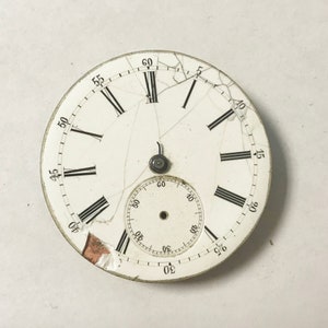 May include: A white pocket watch face with black Roman numeral hour markers and black minute markers. The watch face is cracked and has a smaller circular face inside. The watch face is 55 mm in diameter.
