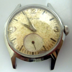 May include: A silver wristwatch with a white face and black markings. The watch has a small second hand dial and the word "ROTON" printed on the face.