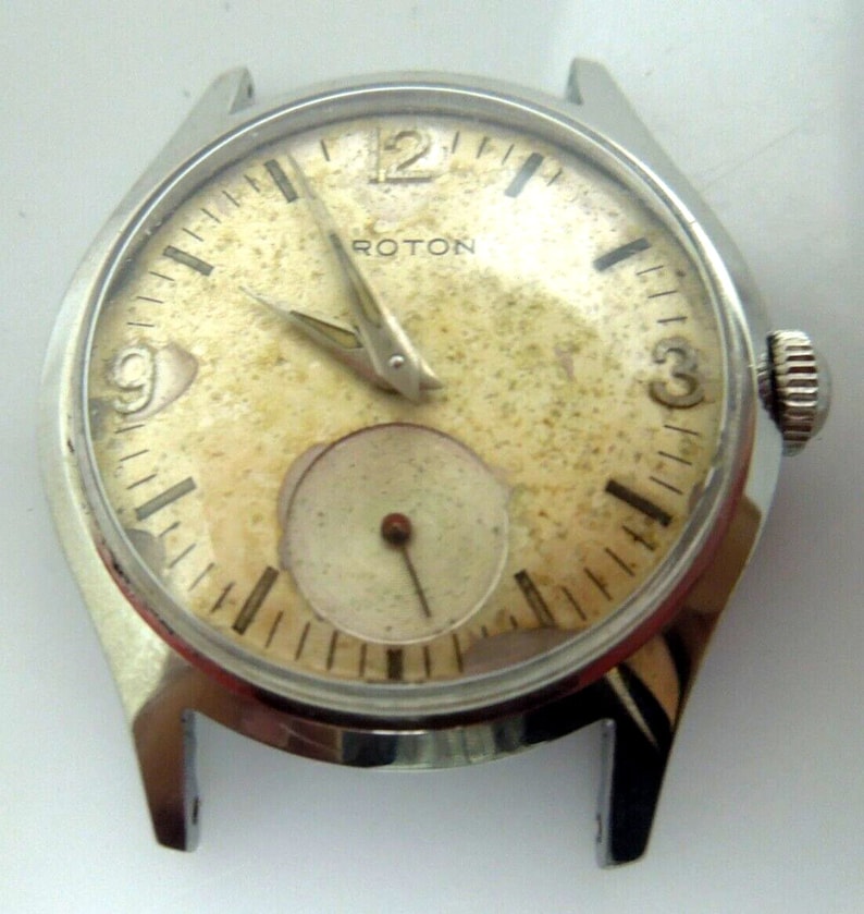 May include: A silver wristwatch with a white face and black markings. The watch has a small second hand dial and the word "ROTON" printed on the face.