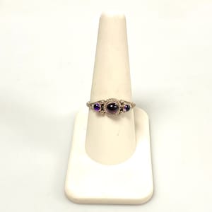 May include: A silver ring with three purple gemstones. The center stone is a larger, round, dark purple gem, flanked by two smaller, oval, lighter purple gems. The ring is displayed on a white jewelry stand.