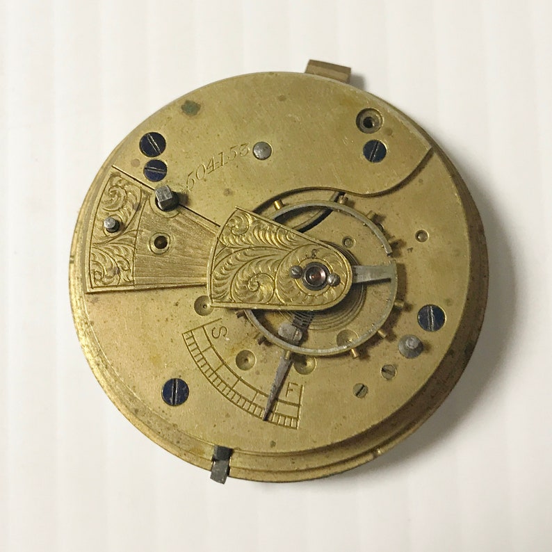 Op de afbeelding: Close-up van een goudkleurig zakhorloge-uurwerk met ingewikkelde gravures en het zichtbare nummer "504756". De wijzerplaat van het horloge ontbreekt.