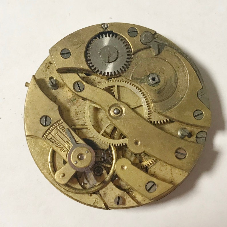 May include: Close-up of a gold-toned pocket watch movement with intricate gears and a lever marked "Alvadec RD".