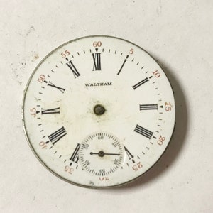 Op de afbeelding: Een wit zakhorloge-horloge met zwarte Romeinse cijfers als uurmarkeringen en zwarte wijzers. De wijzerplaat heeft een kleinere, concentrische cirkel met zwarte markeringen voor seconden. De wijzerplaat is met "WALTHAM" in zwarte letters gemarkeerd.